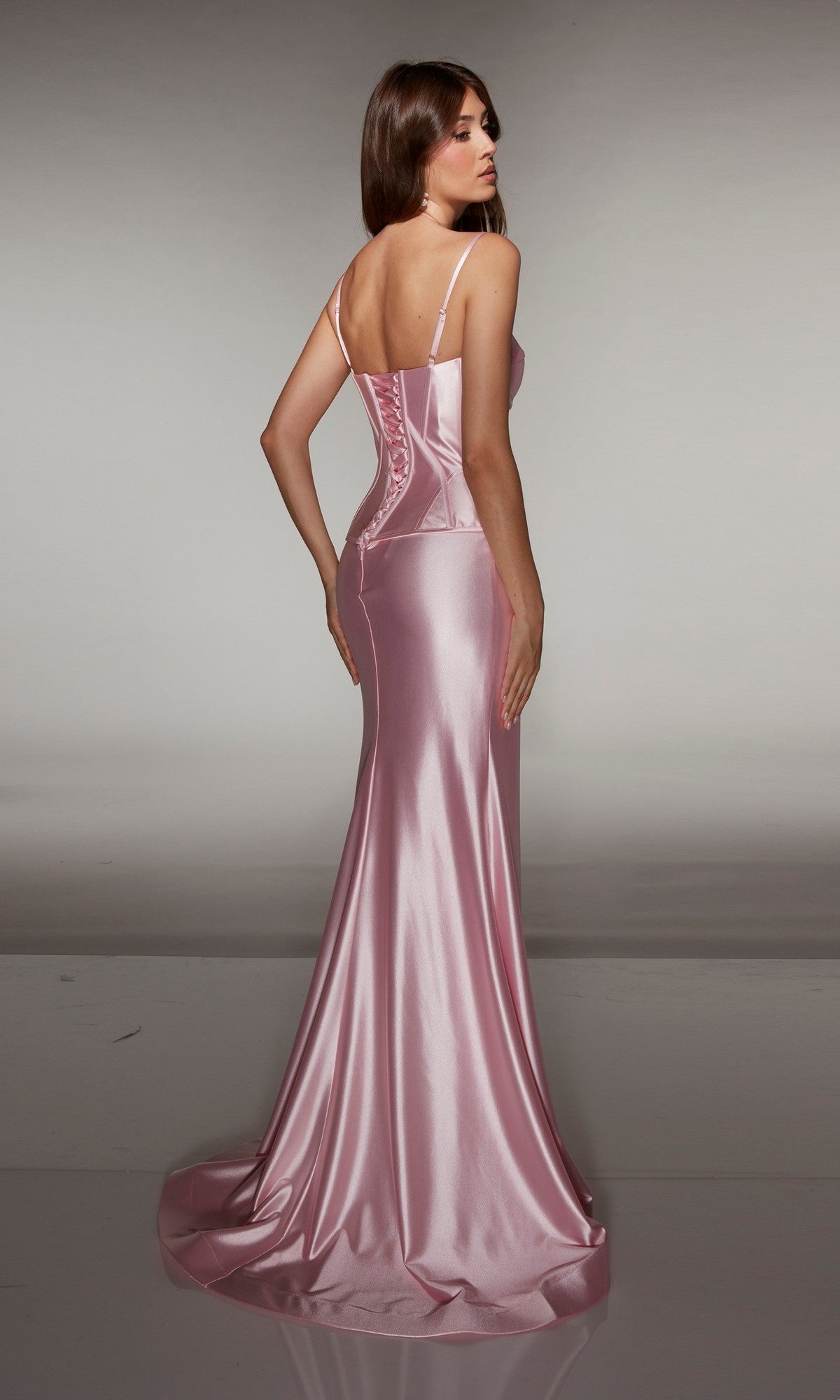 Woman in the Alyce 61983 long prom dress, a floor-length light pink satin gown with a corset bodice and spaghetti straps, shown standing and facing away.